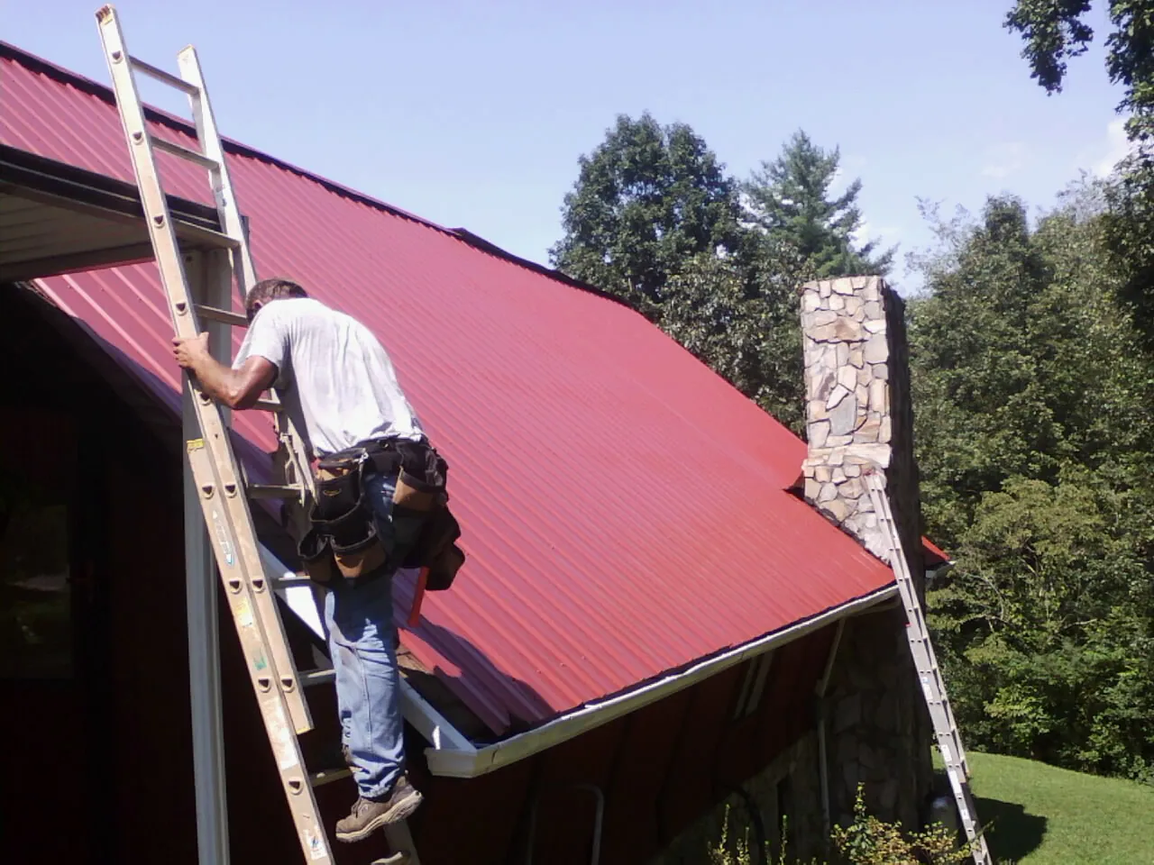 Roofer on a ladder beside a red metal roof and stone chimney in Davidsonville